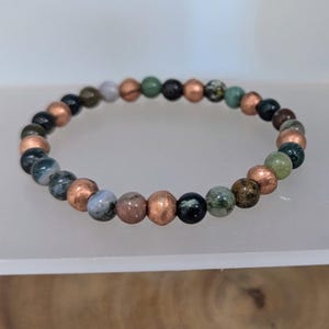 May include: A beaded bracelet featuring a mix of colorful gemstones and copper-colored beads. The gemstones include shades of green, blue, brown, and pink. The bracelet is strung on an elastic band.
