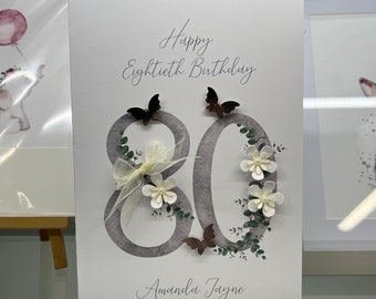 May include: A white card with the text "Happy Eightieth Birthday" and the number 80 decorated with white flowers, green leaves, and brown butterflies. The name "Amanda Jayne" is written at the bottom of the card.