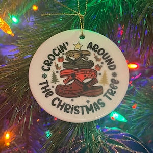 Puede incluir: Adorno navideño de cerámica blanca con una imagen de dibujos animados roja, verde y marrón de una pila de zapatos Crocs. El adorno dice "Crocin' Around The Christmas Tree".