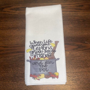 May include: White kitchen towel with a colorful illustration of a pot of food, lemons, and crawfish. The text reads "When life gives you lemons grab some friends and stir the pot."