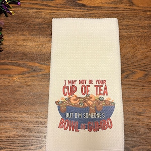 May include: White kitchen towel with a colorful illustration of a bowl of gumbo. The text reads "I may not be your cup of tea, but I'm someone's bowl of gumbo."