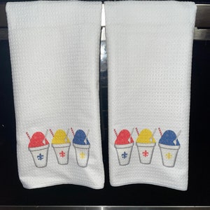 May include: Two white kitchen towels with a row of three colorful snow cone cups embroidered on each towel. The snow cones are red, yellow, and blue.