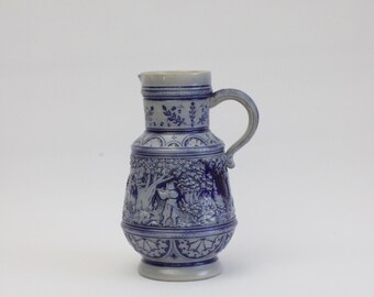 Puede incluir: Una jarra de cerámica azul y blanca con un diseño en relieve de una escena en un bosque. La jarra tiene un asa y un cuerpo redondeado.