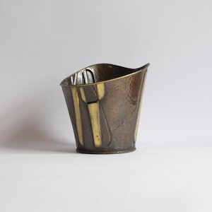 May include: A hammered brass utensil holder with a design of a fork, knife, and spoon. The holder is a dark brown colour with gold accents.