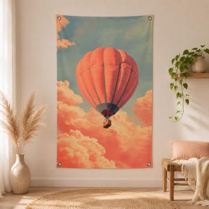 Op de afbeelding: Een wandtapijt met een luchtballon in vintage stijl die door een lucht vol perzikkleurige wolken zweeft. De ballon is koraal van kleur met een grijze basis en een klein mandje. De achtergrond is een verloop van blauw en perzik.