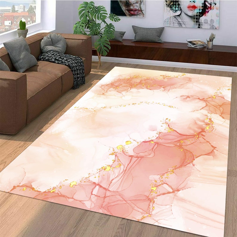 May include: A rectangular rug featuring an abstract design in peach, pink, and white, with gold flecks. The pattern resembles watercolour or ink art, creating a soft, flowing aesthetic. This decorative rug is suitable for living rooms and bedrooms.