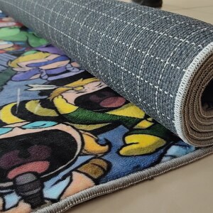 May include: Rolled-up rectangular rug with a cartoon design in bright colours. The rug features a blue, yellow, and green colour scheme. The underside of the rug is grey with a grid pattern, and the edges have a grey border.
