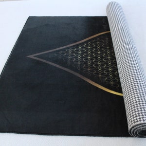 May include: A black prayer rug with a gold geometric design in the center. The rug is rolled up and has a white and black checkered backing.
