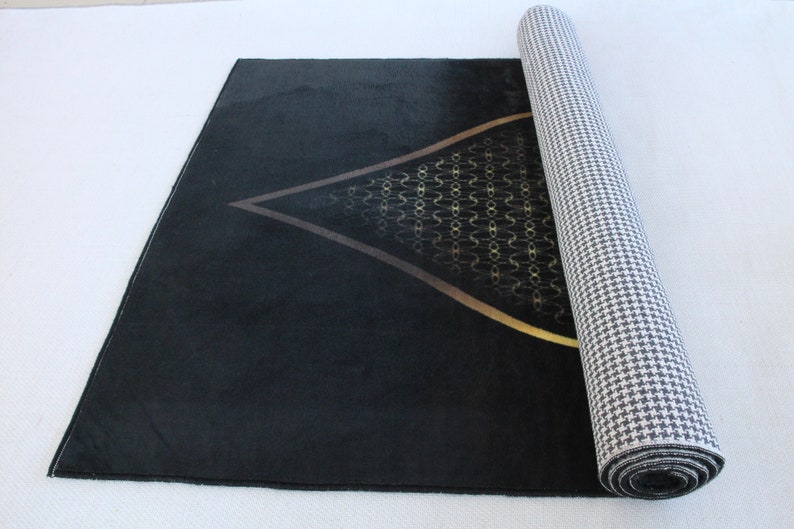 May include: A black prayer rug with a gold geometric design in the center. The rug is rolled up and has a white and black checkered backing.