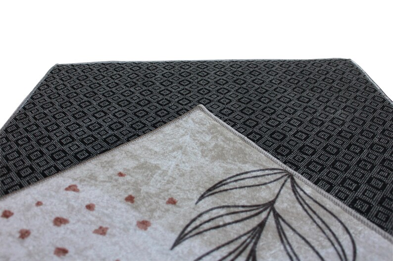 May include: Close-up of a beige rug with a black and white geometric pattern on the reverse. The rug features a floral design with brown leaves and small brown dots.