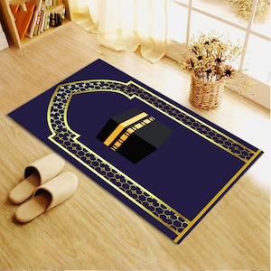 May include: A blue prayer rug with a gold border and a gold arch design. The rug features a black and gold illustration of the Kaaba, a cube-shaped building in Mecca, Saudi Arabia.