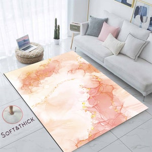 May include: A rectangular rug featuring a watercolour design in peach, pink, and gold hues. The rug has a soft, thick texture, with the words "SOFT & THICK" printed on the lower left corner. The rug is placed on a tiled floor.
