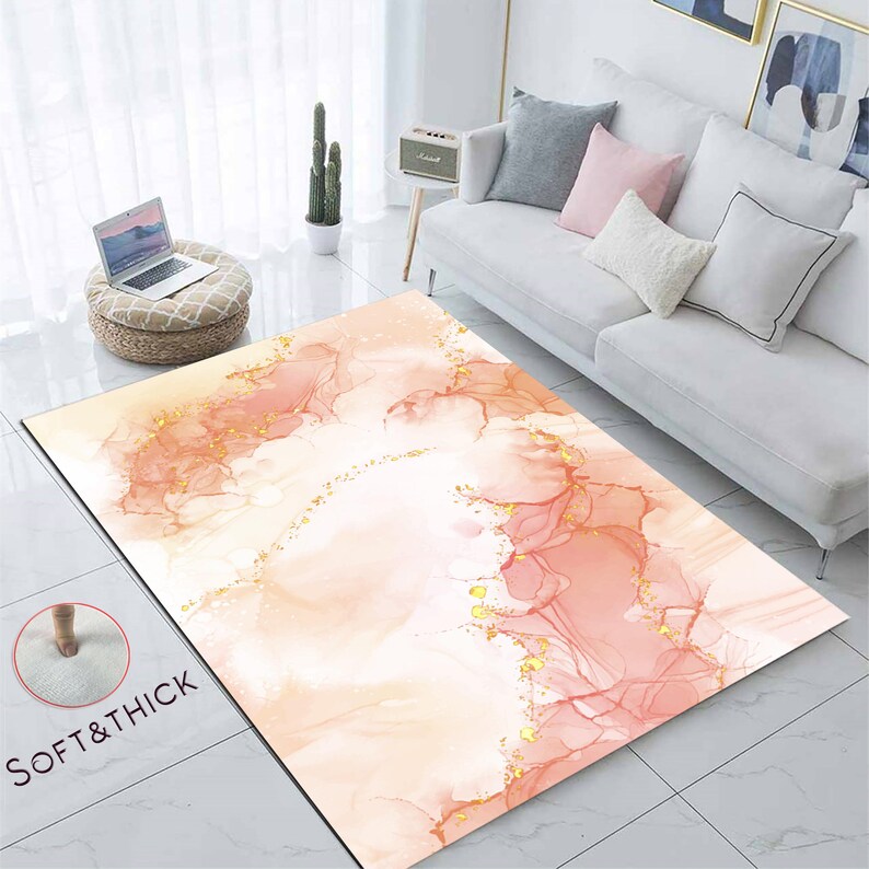 May include: A rectangular rug featuring a watercolour design in peach, pink, and gold hues. The rug has a soft, thick texture, with the words "SOFT & THICK" printed on the lower left corner. The rug is placed on a tiled floor.