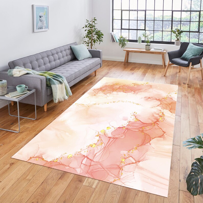May include: A rectangular rug with an abstract design in peach, pink, and gold. The rug has a watercolour-like pattern with flowing lines and gold accents. It is placed on a wooden floor in a living room setting.