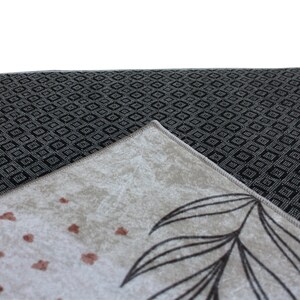 May include: Close-up of a beige rug with a black and white geometric pattern on the back. The rug has a floral design with brown leaves and small brown dots.