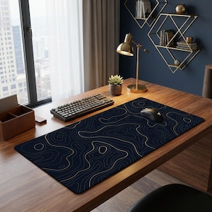 May include: A dark blue desk mat featuring a gold topographic map design. A black keyboard and mouse are on the mat, which is placed on a wooden desk. A gold desk lamp and a small potted plant are also visible. The desk is near a window.
