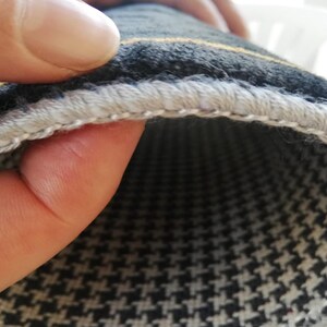 May include: Close-up of a section of a houndstooth patterned fabric. The fabric is black and white with a yellow stripe. The edge is finished with a light gray, stitched border. The fabric is being held by a person's fingers.