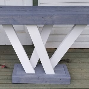 May include: A grey wooden console table with a white painted base. The base is made of four pieces of wood that are arranged in a criss-cross pattern.