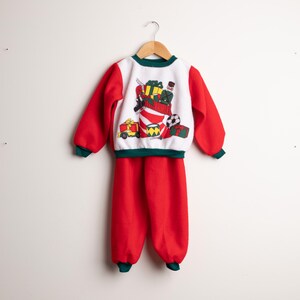 May include: A festive two-piece Christmas outfit. The top is white with a red and green design, showing a stocking filled with presents. The sleeves and trousers are red with green trim. The outfit is hanging on a wooden hanger.