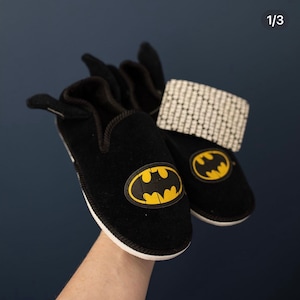 May include: A pair of black fleece slippers with a yellow Batman logo on each slipper. The slippers have a white trim around the edges.