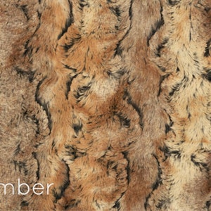 K&ouml;nnte beinhalten: Nahaufnahme eines Kunstpelzstoffes mit einem braunen, schwarzen und beigefarbenen Farbmuster. Der Stoff hat ein wellenf&ouml;rmiges Muster und eine weiche, pl&uuml;schige Textur. "Amber" ist in wei&szlig;er Schrift am unteren Rand des Bildes geschrieben.