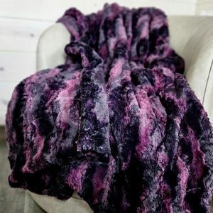 May include: A plush, faux fur blanket in shades of purple, black, and gray is draped over a light-colored armchair. The blanket's texture is soft and fluffy, with a marbled appearance. The color palette creates a luxurious and cozy feel.
