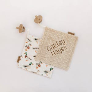 Puede incluir: Un juego de mantas de confort para bebés con un estampado de patos y un chupete. Una manta es beige con un estampado de patos, la otra es beige con el nombre "Oakley Hayes" bordado. El chupete está sujeto con una cuerda blanca.