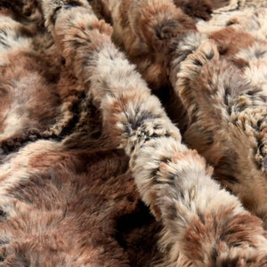 May include: Close-up of a brown, gray, and white faux fur fabric. The fabric has a soft, fluffy texture and a swirling pattern.