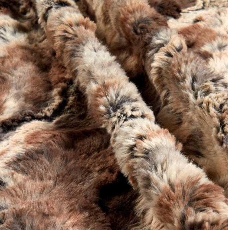 May include: Close-up of a brown, gray, and white faux fur fabric. The fabric has a soft, fluffy texture and a swirling pattern.
