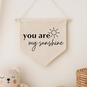 Op de afbeelding: Een witte canvasbanner met zwarte tekst die "you are my sunshine" luidt en een eenvoudige zonafbeelding. De banner hangt aan een witte muur met een houten stok aan de bovenkant.