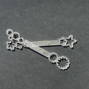 May include: Two clear plastic bubble wands with names "Elena" and "Indigo" engraved on them. The wands have different designs, one with stars and the other with circles.