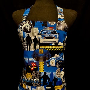 May include: A blue and white apron with a police theme. The apron features images of police cars, officers, and police line do not cross signs.