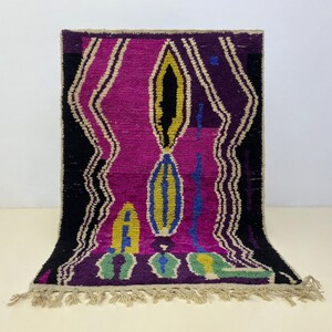 Peut inclure: Un tapis rectangulaire avec un motif abstrait. Le tapis présente un champ central magenta avec des bordures noires et des accents jaunes, bleus et verts. Le tapis a une frange le long du bord inférieur.
