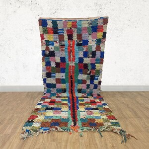 Peut inclure: Un tapis marocain coloré avec un motif géométrique dans des tons de bleu, rouge, vert, jaune et blanc. Le tapis a une bordure à franges et est fait de laine.