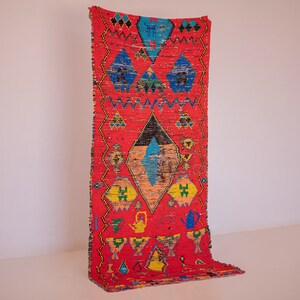 May include: A bright red rectangular rug with geometric patterns in blue, yellow, black, and orange. The rug features diamond and triangle shapes, and decorative elements. The rug is displayed against a white wall.