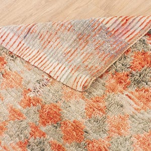 May include: A hand-woven rug with a textured, fluffy surface. The rug features a pattern of orange and beige stripes with a subtle floral design. The rug is folded in half, revealing the underside, which is a solid beige color.