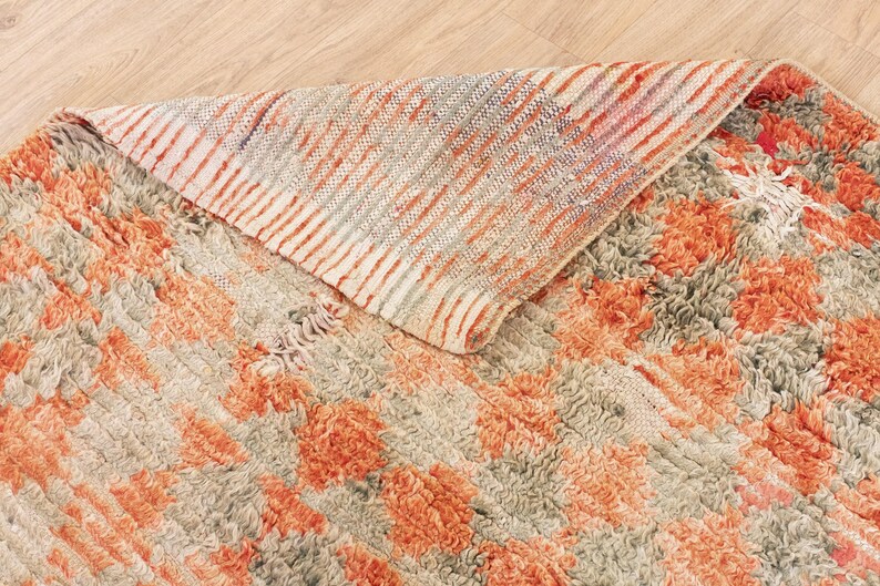 May include: A hand-woven rug with a textured, fluffy surface. The rug features a pattern of orange and beige stripes with a subtle floral design. The rug is folded in half, revealing the underside, which is a solid beige color.