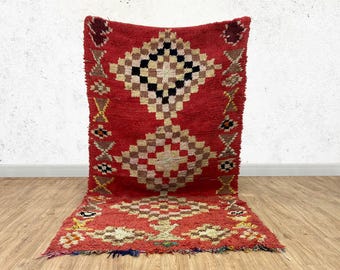 May include: A red, handwoven Moroccan rug with a geometric pattern in shades of brown, black, and white. The rug has a fringed edge and is made of wool.