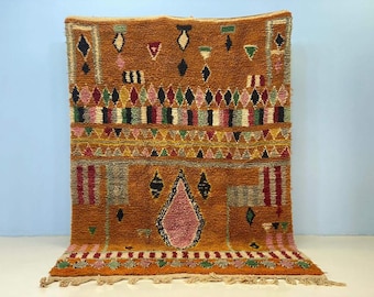 Puede incluir: Una alfombra rectangular tejida a mano con un fondo naranja cálido. La alfombra presenta patrones geométricos en negro, rosa, verde y amarillo. El diseño incluye formas de diamante y triángulo, con un borde con flecos en la parte inferior. La alfombra es para la decoración del hogar.