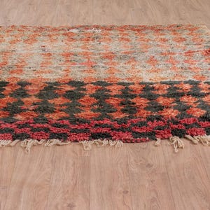 May include: A hand-woven rug with a geometric pattern in shades of red, orange, black, and white. The rug has a fringe border and is made of wool.