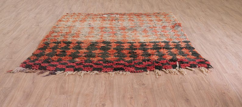 May include: A hand-woven rug with a geometric pattern in shades of red, orange, black, and white. The rug has a fringe border and is made of wool.