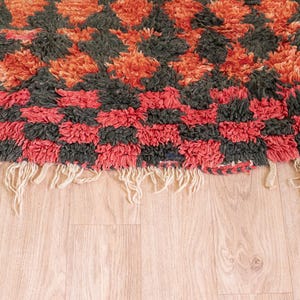 May include: Close-up of a red, black, and orange patterned rug with a fringe border. The rug is made of a thick, fluffy material.