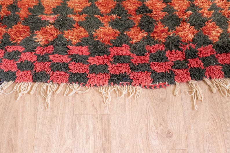 May include: Close-up of a red, black, and orange patterned rug with a fringe border. The rug is made of a thick, fluffy material.