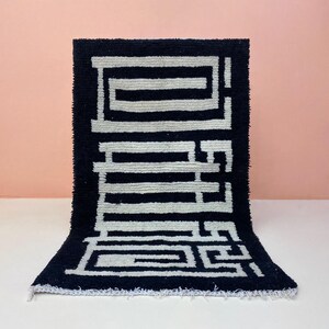 Peut inclure: Tapis rectangulaire bleu foncé avec un motif géométrique blanc. Le tapis est posé sur une surface blanche avec un fond rose. Le motif est composé de formes et de lignes interconnectées, créant un design abstrait.