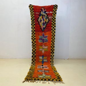 May include: A vibrant, rectangular runner rug with a geometric design. The rug features a central orange field with colorful star-like patterns and a red top section. The edges are bordered with a black and yellow checkered pattern, and the rug has fringe.