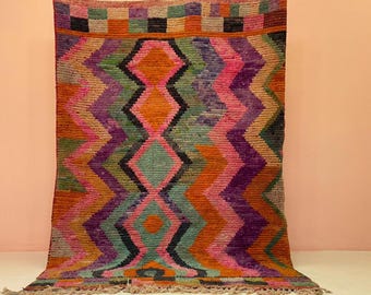 May include: A vibrantly colored, hand-woven rug with a geometric design. The rug features a repeating pattern of zigzags and diamond shapes in shades of orange, pink, purple, green, and black. The rug has a fringed edge and a textured surface.