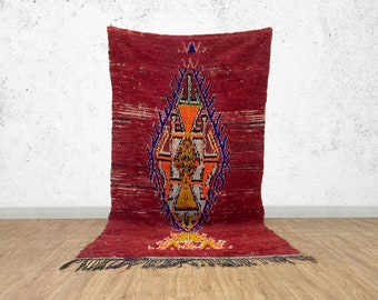 May include: A red Moroccan rug with a geometric pattern in shades of blue, orange, and yellow. The rug has a fringe border.