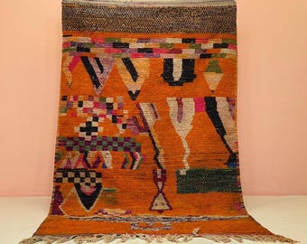 Puede incluir: Alfombra marroquí naranja con diseños geométricos en negro, blanco, rosa y verde. La alfombra tiene una superficie texturizada y un borde con flecos. El diseño incluye triángulos, cuadrados y formas abstractas, creando un textil vibrante y artístico.