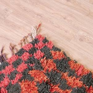 May include: A close-up of a colorful rug with a checkered pattern in shades of red, orange, and black. The rug has a fluffy texture and is made of wool.
