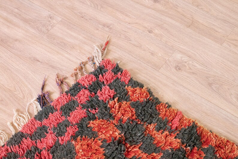 May include: A close-up of a colorful rug with a checkered pattern in shades of red, orange, and black. The rug has a fluffy texture and is made of wool.